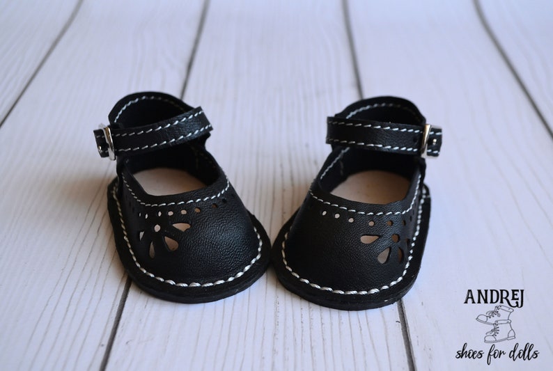 May include: A pair of black leather doll shoes with white stitching and a buckle closure. The shoes have a cut-out design and are made for dolls.