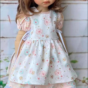 May include: A porcelain doll wearing a white dress with pink and blue floral print, a pink bow in her hair, and white shoes. The doll is standing on a green grassy surface.