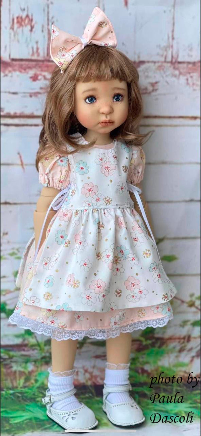 May include: A porcelain doll wearing a white dress with pink and blue floral print, a pink bow in her hair, and white shoes. The doll is standing on a green grassy surface.