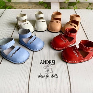 May include: A set of ten miniature leather shoes for dolls in various colors, including blue, red, white, and beige. The shoes have a cut-out design and are all closed-toe. The shoes are arranged on a wooden surface. The text "ANDRJEJ shoes for dolls" is visible in the image.