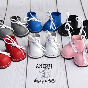 Puede incluir: Un conjunto de diez zapatos de muñeca de cuero en miniatura en varios colores, incluyendo rojo, blanco, azul, negro, gris y rosa. Los zapatos tienen cordones blancos y están dispuestos en pares sobre una superficie de madera blanca. El texto "ANDRJEJ shoes for dolls" está impreso debajo de los zapatos.