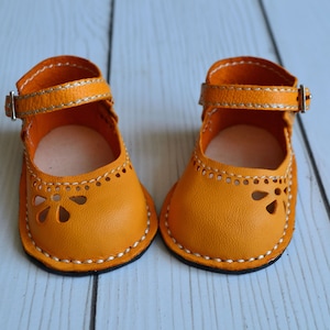 May include: A pair of yellow leather doll shoes with white stitching and a buckle closure. The shoes have a cut-out design on the toe.