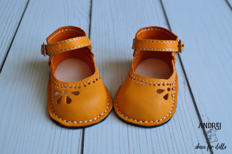 May include: A pair of yellow leather doll shoes with white stitching and a buckle closure. The shoes have a cut-out design on the toe.
