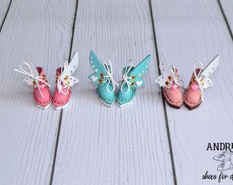 Könnte beinhalten: Drei Paar Miniatur-Puppenschuhe aus Leder mit Schmetterlingsflügeln. Die Schuhe sind rosa, türkis und rosa mit weißen Flügeln. Die Schuhe haben Schnürsenkel und sind mit goldenen Akzenten verziert.