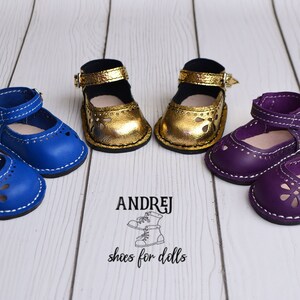 May include: Three pairs of doll shoes in blue, gold, and purple. Each pair has a buckle and a cut-out design. The shoes are on a white wooden surface. The text "ANDRJEJ shoes for dolls" is visible below the shoes.
