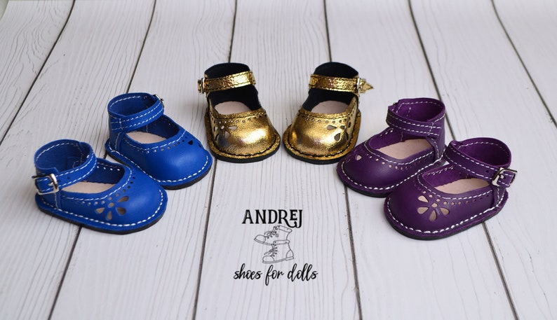 May include: Three pairs of doll shoes in blue, gold, and purple. Each pair has a buckle and a cut-out design. The shoes are on a white wooden surface. The text "ANDRJEJ shoes for dolls" is visible below the shoes.