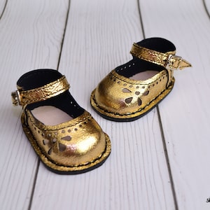 May include: A pair of gold leather doll shoes with cut-out designs and black straps. The shoes have a buckle closure and are on a white wooden surface.