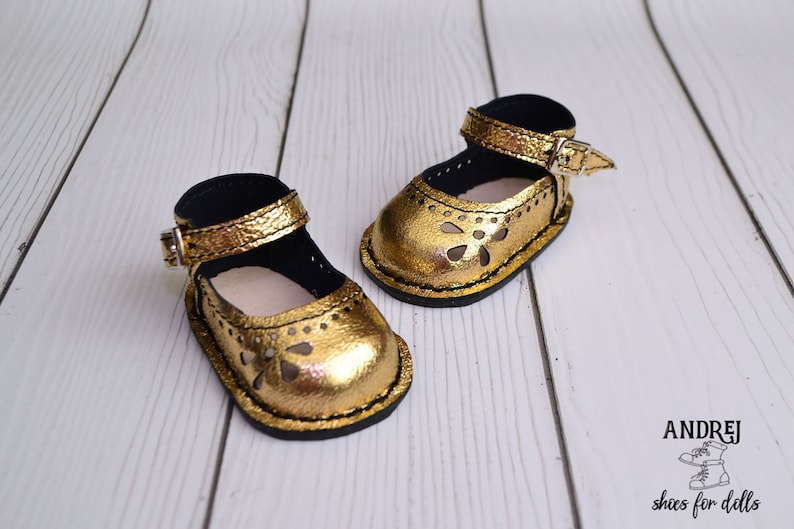 May include: A pair of gold leather doll shoes with cut-out designs and black straps. The shoes have a buckle closure and are on a white wooden surface.