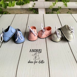 May include: Four pairs of miniature leather shoes for dolls. Two pairs are light blue, one pair is pink, and one pair is white. The shoes have white laces and are arranged on a wooden surface. The text "ANDRJEJ shoes for dolls" is printed below the shoes.
