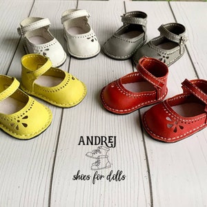 May include: Six pairs of miniature leather shoes for dolls in yellow, white, gray, and red. The shoes have a strap with a buckle and a flower-shaped cut-out design. The shoes are arranged on a wooden surface.  ANDREJ shoes for dolls.