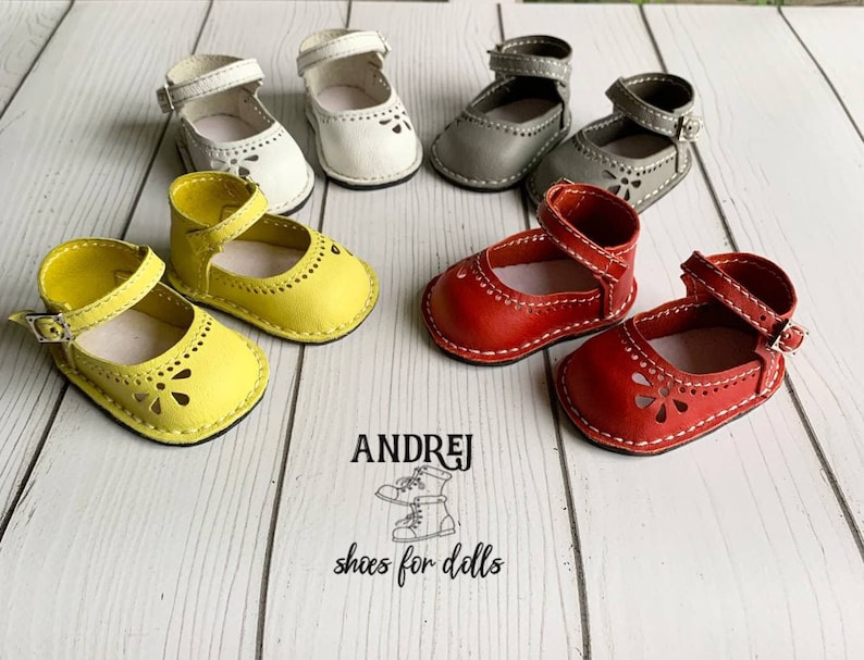 May include: Six pairs of miniature leather shoes for dolls in yellow, white, gray, and red. The shoes have a strap with a buckle and a flower-shaped cut-out design. The shoes are arranged on a wooden surface.  ANDREJ shoes for dolls.