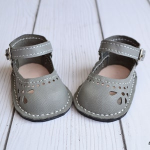 May include: A pair of gray leather doll shoes with ankle straps and cut-out detailing. The shoes have white stitching and are on a white wooden surface.