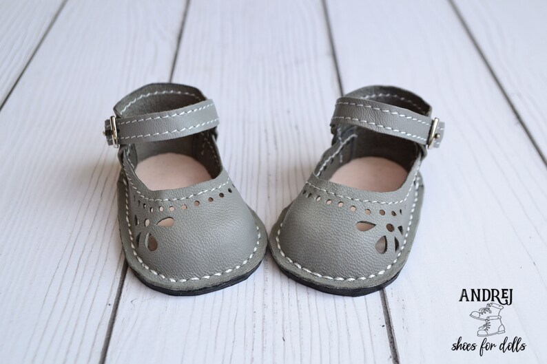 May include: A pair of gray leather doll shoes with ankle straps and cut-out detailing. The shoes have white stitching and are on a white wooden surface.