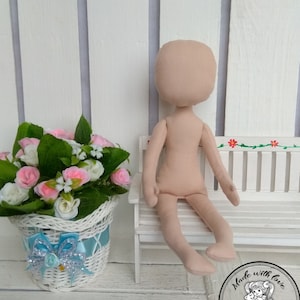 May include: Unpainted doll made of beige fabric sitting on a white bench. The doll has a round head, arms, and legs. The doll is sitting with its legs crossed. The doll is in front of a white wooden wall. A white wicker basket with a blue bow is in the foreground. The basket is filled with pink and white flowers.