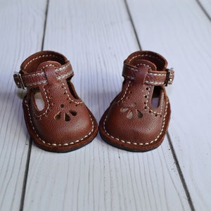 May include: A pair of miniature brown leather doll shoes with a T-strap design. The shoes feature decorative cutouts, white stitching, and small buckles. The soles are rounded, and the shoes are set against a light wood background.