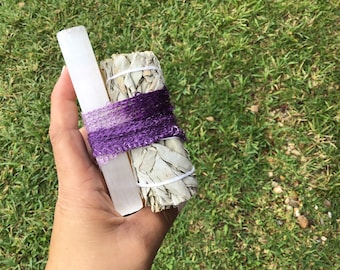 May include: A bundle of white sage smudge sticks tied with purple string, next to a white selenite stick.