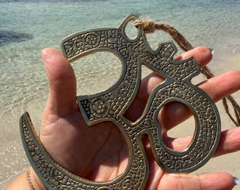 May include: A brass-colored Om symbol pendant with intricate floral patterns. The pendant is suspended by a natural fiber cord. The image is taken outdoors, with a beach and ocean in the background.