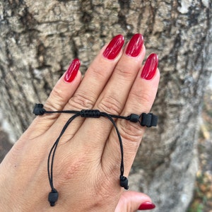 May include: A black adjustable bracelet with black beads. The bracelet is tied around a person's wrist.