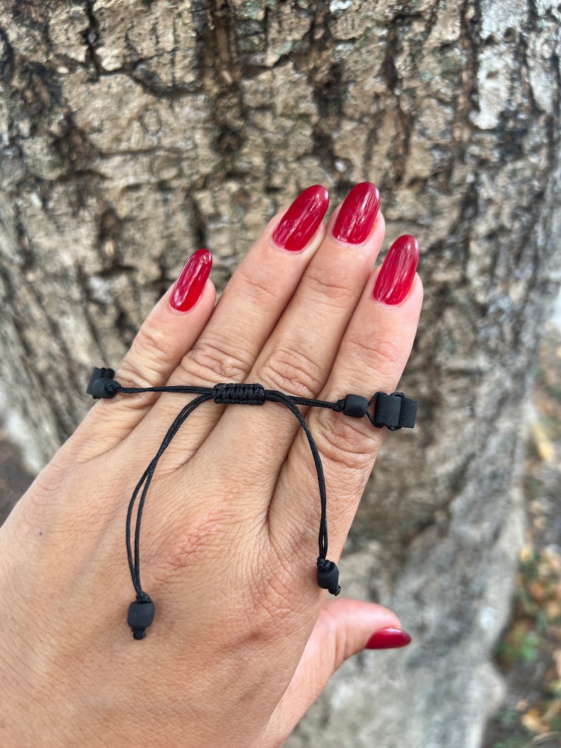 May include: A black adjustable bracelet with black beads. The bracelet is tied around a person's wrist.