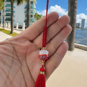 May include: A red decorative hanging charm featuring a small white ceramic cat bead with black and red floral details, accented by gold-toned metal beads, a braided red cord, and a long red fabric tassel at the bottom for ornamental display.