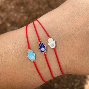 May include: Three red string bracelets with gold hamsa hand charms. Each charm has a blue, white, or turquoise enamel finish and a clear rhinestone in the center.