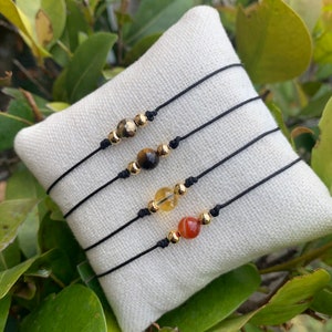 May include: Four black string bracelets with gold beads and different colored gemstone beads. The bracelets are displayed on a white fabric surface.