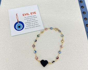 Puede incluir: Un brazalete de cadena dorada con un dije de corazón negro y múltiples dijes de ojo turco de colores. El brazalete está sobre una superficie blanca con una tarjeta que dice "Evil Eye Its watchful gaze is believed to keep us safe from jealousy and harm, so they're given to bless & protect those we truly admire."