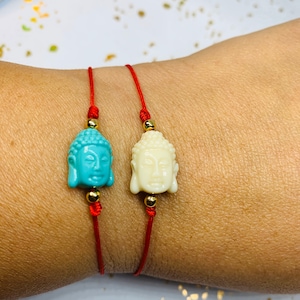 May include: Two red string bracelets with gold beads and a Buddha head charm. One charm is turquoise blue and the other is white.