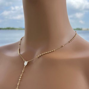 Puede incluir: Un collar de rosario dorado con un peque&ntilde;o dije de cruz y un dije que representa a la Virgen Mar&iacute;a. El collar se muestra en un maniqu&iacute;.