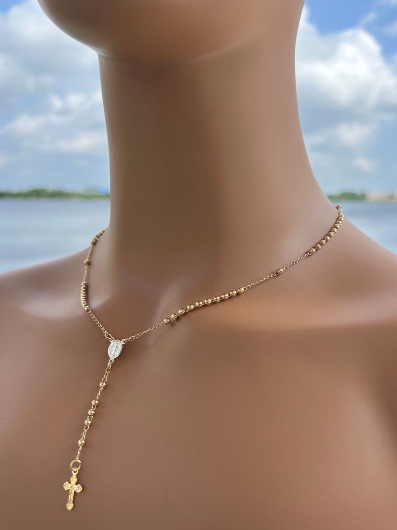 Puede incluir: Un collar de rosario dorado con un peque&ntilde;o dije de cruz y un dije que representa a la Virgen Mar&iacute;a. El collar se muestra en un maniqu&iacute;.