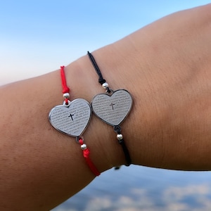Puede incluir: Dos pulseras en forma de corazón con una inscripción religiosa en español. Las pulseras están hechas de cuerda negra y roja y presentan un dije de corazón plateado con una cruz. Las pulseras se llevan en la muñeca.