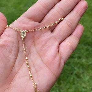 Puede incluir: Un collar de cadena de oro con un peque&ntilde;o colgante de cruz y un dije redondo con una imagen religiosa. El collar est&aacute; en una mano.