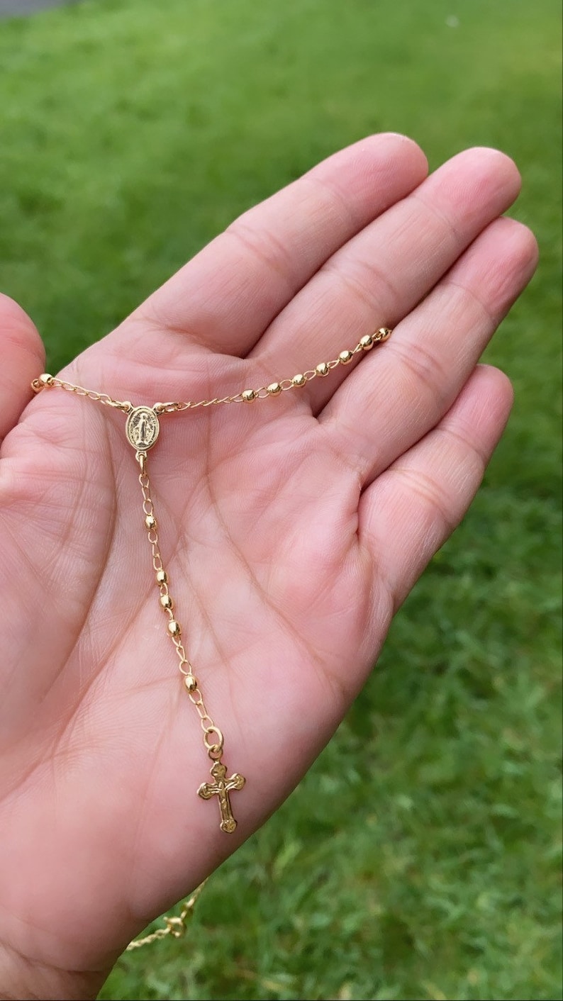 Puede incluir: Un collar de cadena de oro con un peque&ntilde;o colgante de cruz y un dije redondo con una imagen religiosa. El collar est&aacute; en una mano.