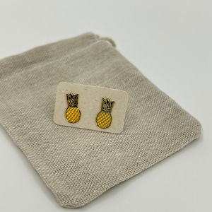 May include: A pair of gold pineapple stud earrings on a small card. The pineapples are yellow with black outlines.