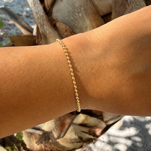 May include: A delicate gold beaded bracelet. The bracelet is composed of small, evenly spaced, spherical beads. The jewelry is worn on a wrist, with a natural background of a palm tree and sunlight.