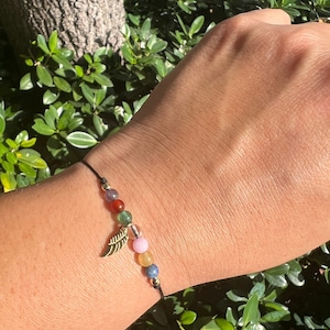 Puede incluir: Un brazalete de cuerda negra con un dije de hoja de oro y cuentas de piedras preciosas de colores. Las cuentas son rojas, verdes, transparentes, rosas, amarillas y azules.