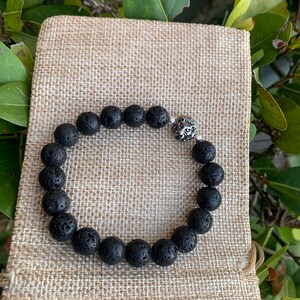 Op de afbeelding: Zwarte lavastenen armband met een zilveren leeuwenkop bedel. De armband wordt getoond op een bruine jute zak.