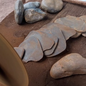 May include: A close-up of a pile of smooth, gray-brown stones, some of which are stacked on top of each other. The stones are on a wooden surface, and there is a metal disc in the foreground.