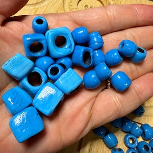 May include: A collection of bright sky blue beads in various shapes, including cubes, spheres, and rounded forms. The beads have a smooth, glossy finish with subtle white marbling throughout, and each piece features a central hole for stringing.