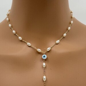 May include: Gold-toned necklace featuring a strand of small, oval pearls. A blue and white evil eye charm hangs below the pearl strand, with a dangling chain of pearls ending in a gold Hamsa hand charm. The necklace is displayed on a person.