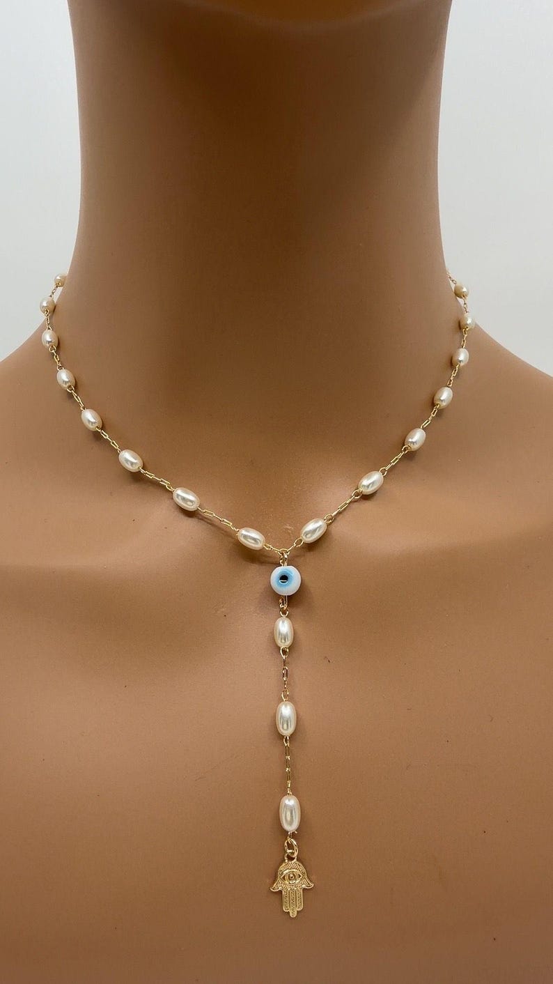 May include: Gold-toned necklace featuring a strand of small, oval pearls. A blue and white evil eye charm hangs below the pearl strand, with a dangling chain of pearls ending in a gold Hamsa hand charm. The necklace is displayed on a person.