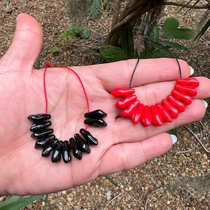 May include: Two necklaces with teardrop-shaped beads. One necklace features glossy black beads strung on a red cord. The other necklace has bright red beads on a black cord. Both necklaces are displayed on a hand.