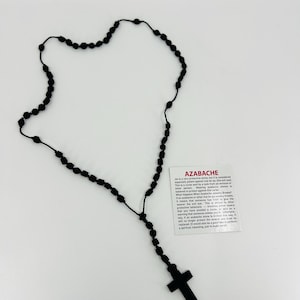 May include: Black beaded rosary necklace with a wooden cross pendant. The necklace is accompanied by a small card with the text "AZABACHE" and information about the stone.