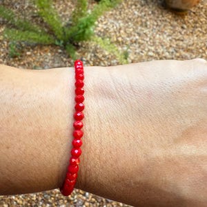 May include: A red beaded bracelet is worn on a person's wrist. The bracelet is made of small, round, faceted beads. The beads are a vibrant red color and are strung together to form a continuous loop. The bracelet is a simple, elegant piece of jewelry.