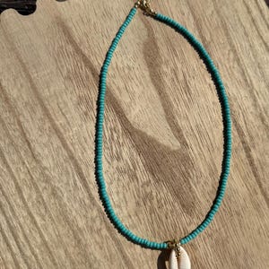 May include: A turquoise beaded necklace with a white seashell pendant. The necklace is on a wooden surface.