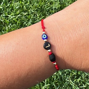 May include: Red string bracelet with a blue evil eye charm, a black bead with the letter "L" in gold lettering, and a black bead.