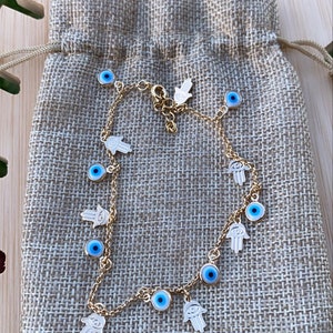 Puede incluir: Un brazalete de cadena dorada con dijes de ojo turco azul y mano de hamsa. El brazalete está en una bolsa de arpillera marrón.