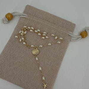May include: A gold-colored necklace with a seashell pendant and a small starfish charm, adorned with white pearls. The necklace rests on a beige drawstring pouch with wooden beads on the drawstrings. The jewelry is likely for a beach or nautical theme.