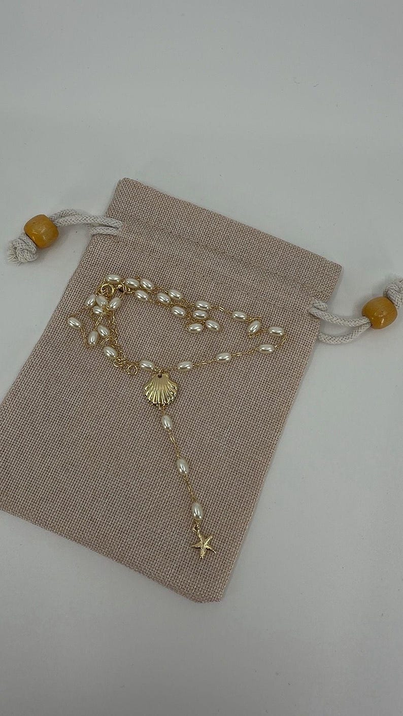 May include: A gold-colored necklace with a seashell pendant and a small starfish charm, adorned with white pearls. The necklace rests on a beige drawstring pouch with wooden beads on the drawstrings. The jewelry is likely for a beach or nautical theme.