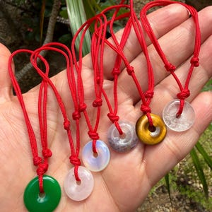 Puede incluir: Una colección de seis colgantes de piedras preciosas con cordones rojos. Los colgantes son de varios colores, incluyendo verde, rosa, blanco y dorado. Cada colgante tiene un agujero en el centro. Los colgantes se muestran en una mano.